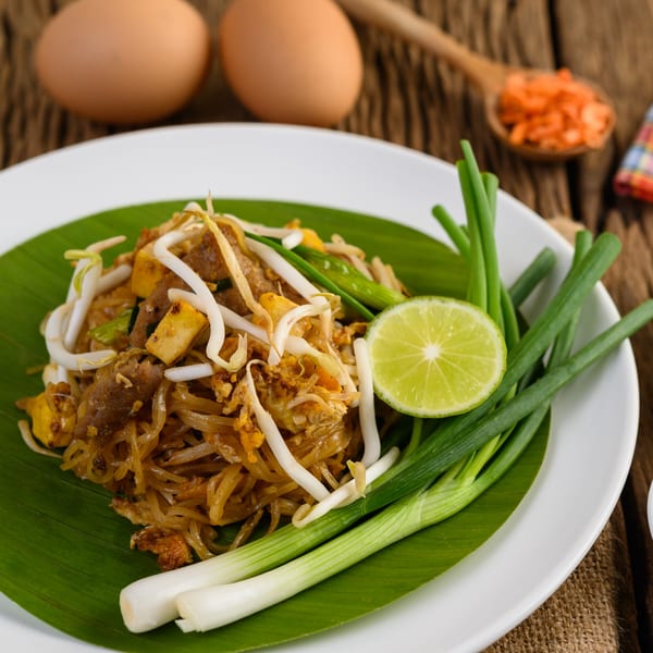 Pad Thai noodles with bean sprouts, tofu, meat, lime, and green onions on a banana leaf.